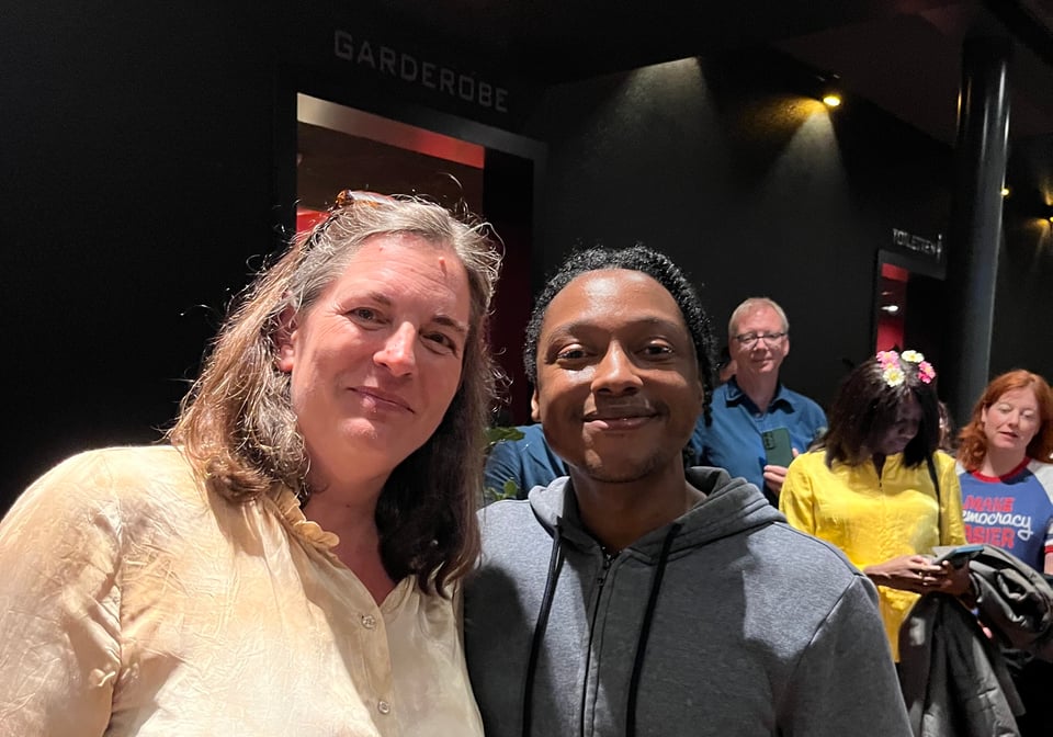 A smiling white woman in a gold shirt stands next to a smiling black man in a grey hoodie.
