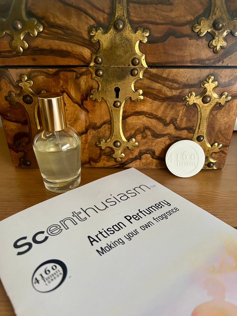 A small bottle of cloudy perfume is in front of a wooden treasure chest by a booklet with 4160 Tuesdays branding on.