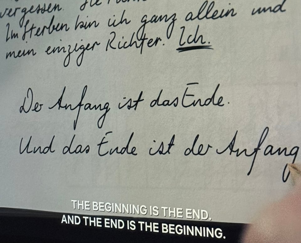 A hand writing a letter that says “The beginning is the end, and the end is the beginning.“ in German.