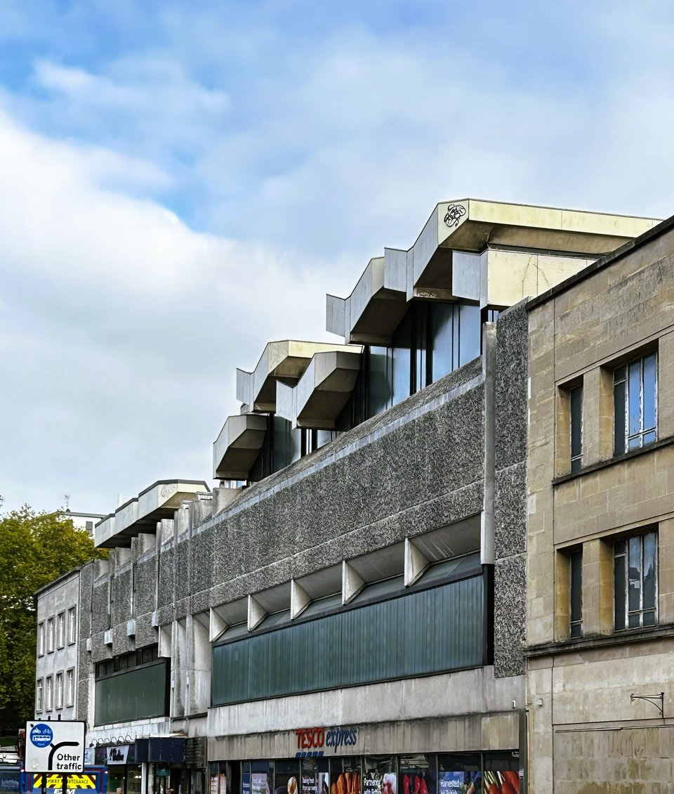 A brutalist building beneath a pale blue sky with high white clouds. The building's first floor is a wall of thick vertical glass blocks. Above that is a wall of concrete and above that is more glass, surmounted by concrete roof beams that have been designed to look like huge white doves. On the ground floor is a Tesco Express.