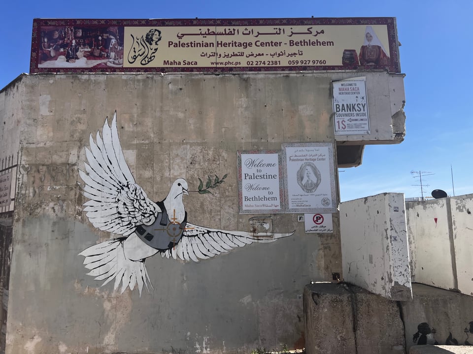 The side of a concrete building with a painting of a dove, and a sign above the wall that reads "Palestinian Heritage Center - Bethlehem" in English, and with Arabic writing as well