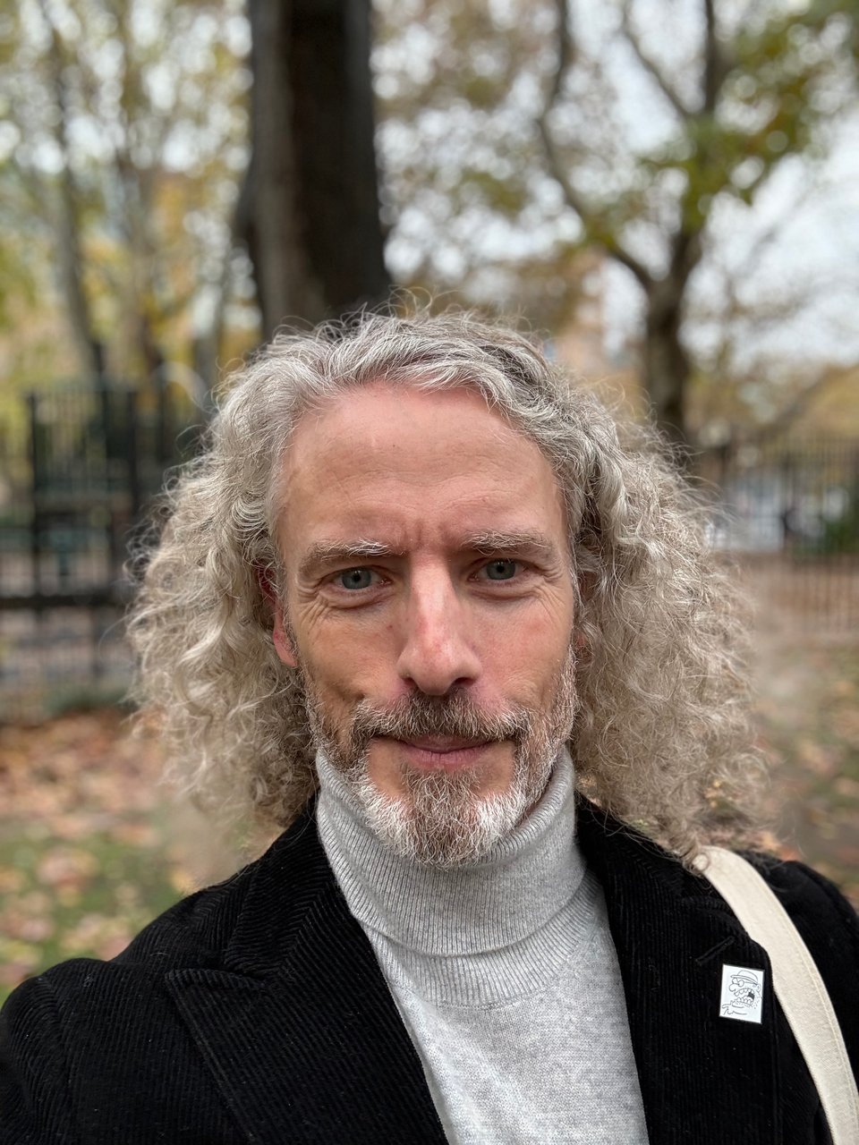 selfie of white man in black corduroy suit and silver-gray turtleneck, with long hair, standing in a park in autumn