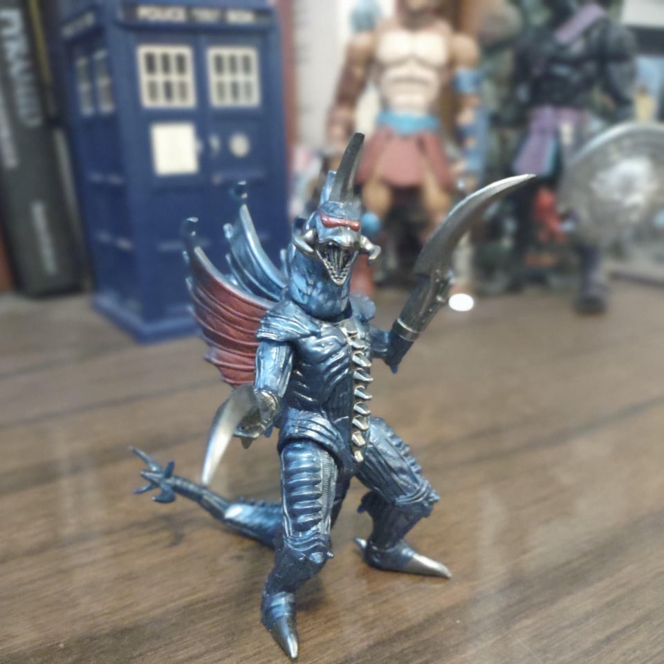 A tiny kaiju action figure stands on a wooden desk. He has one wide red eye, almost a visor, large blades for hands, and three wing-like fins sticking out from his back.