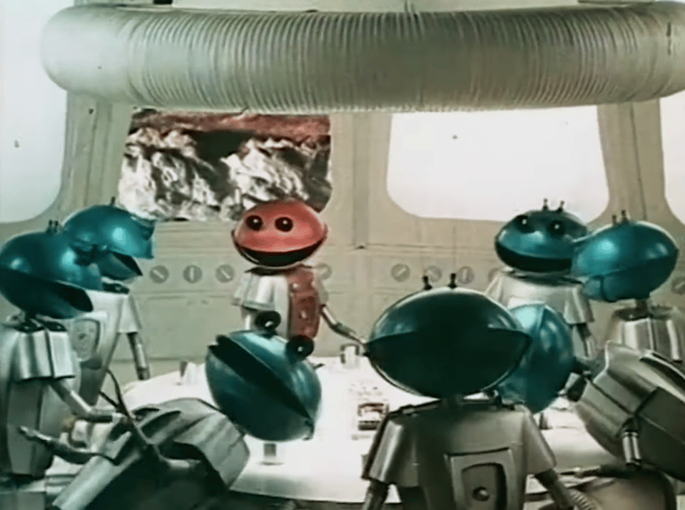 A screenshot from a Cadbury's Smash (instant mashed potato) television advert. A group of simplistic model Martians are laughing round a table. One of them has fallen over during the take.