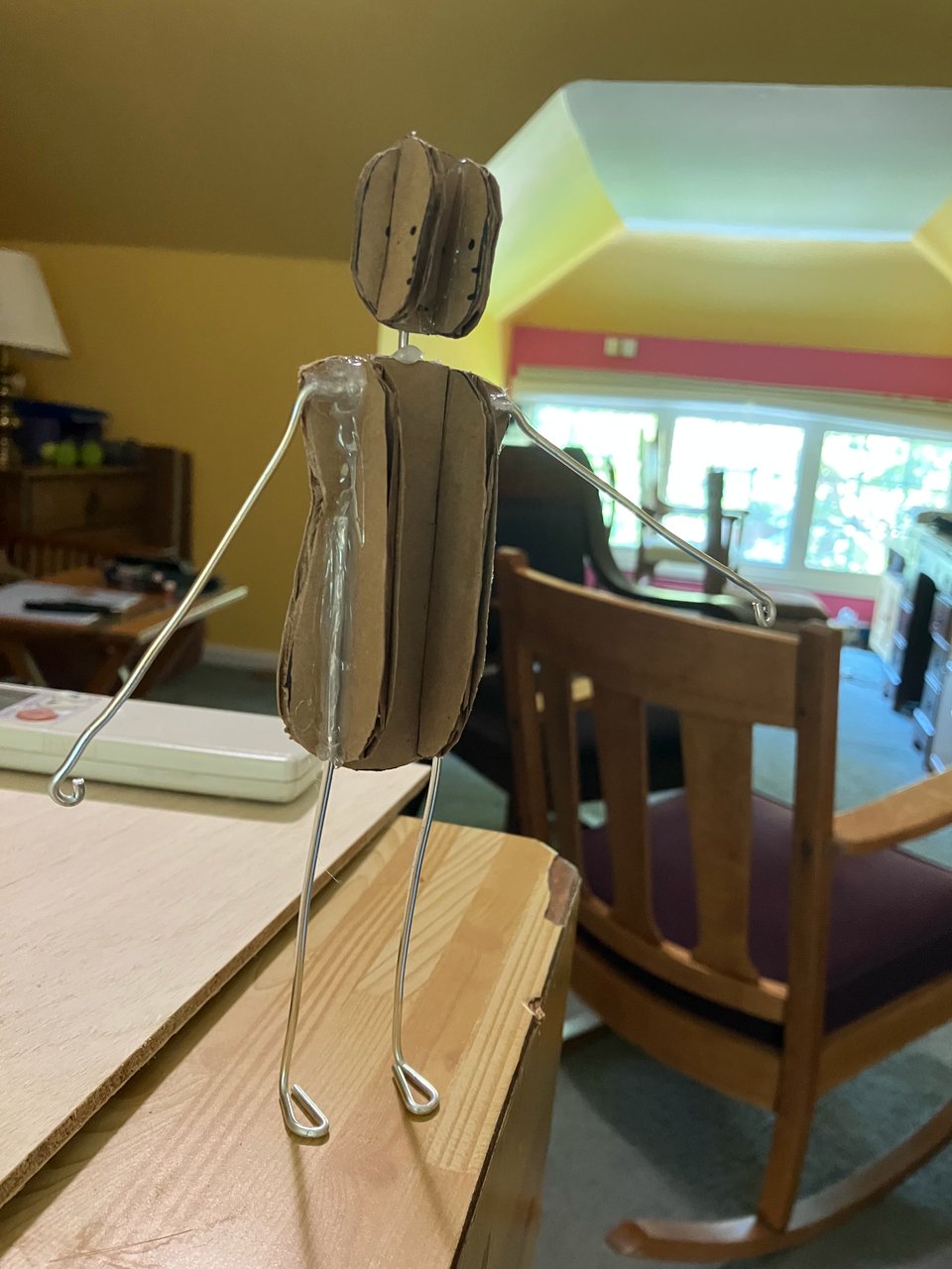 A humanoid puppet with cardboard pieces intersecting each other for roundness for the body and head, and wire limbs with loops for hands and feet hot-glued to the cardboard body.