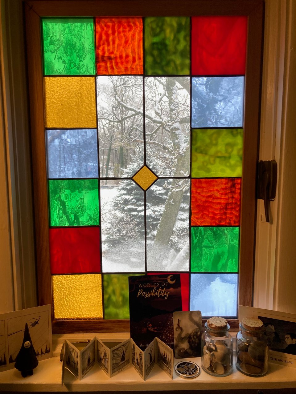 A view through a stained-glass window overlooking snow-dusted trees and branches. At the base of the window is a sill covered with ephemera: a Jon Carling
