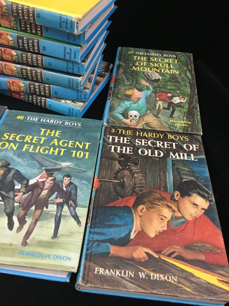 A pile of The Hardy Boys hardcover books, with three lying flat on the table: The Secret of Skull Mountain (book 27), The Secret of the Old Mill (book 3) and The Secret Agent on Flight 101 (book 46).