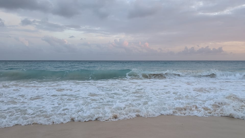 A glass-green and white foamy ocean, waves crashing toward the camera. The sky is full of downy grey clouds.