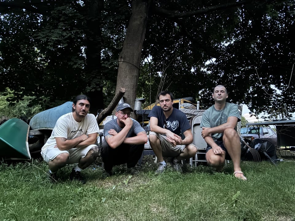 Four men crouch on grass in front of a turned-over canoe, a tree and a metal folding chair in the upright and locked position.