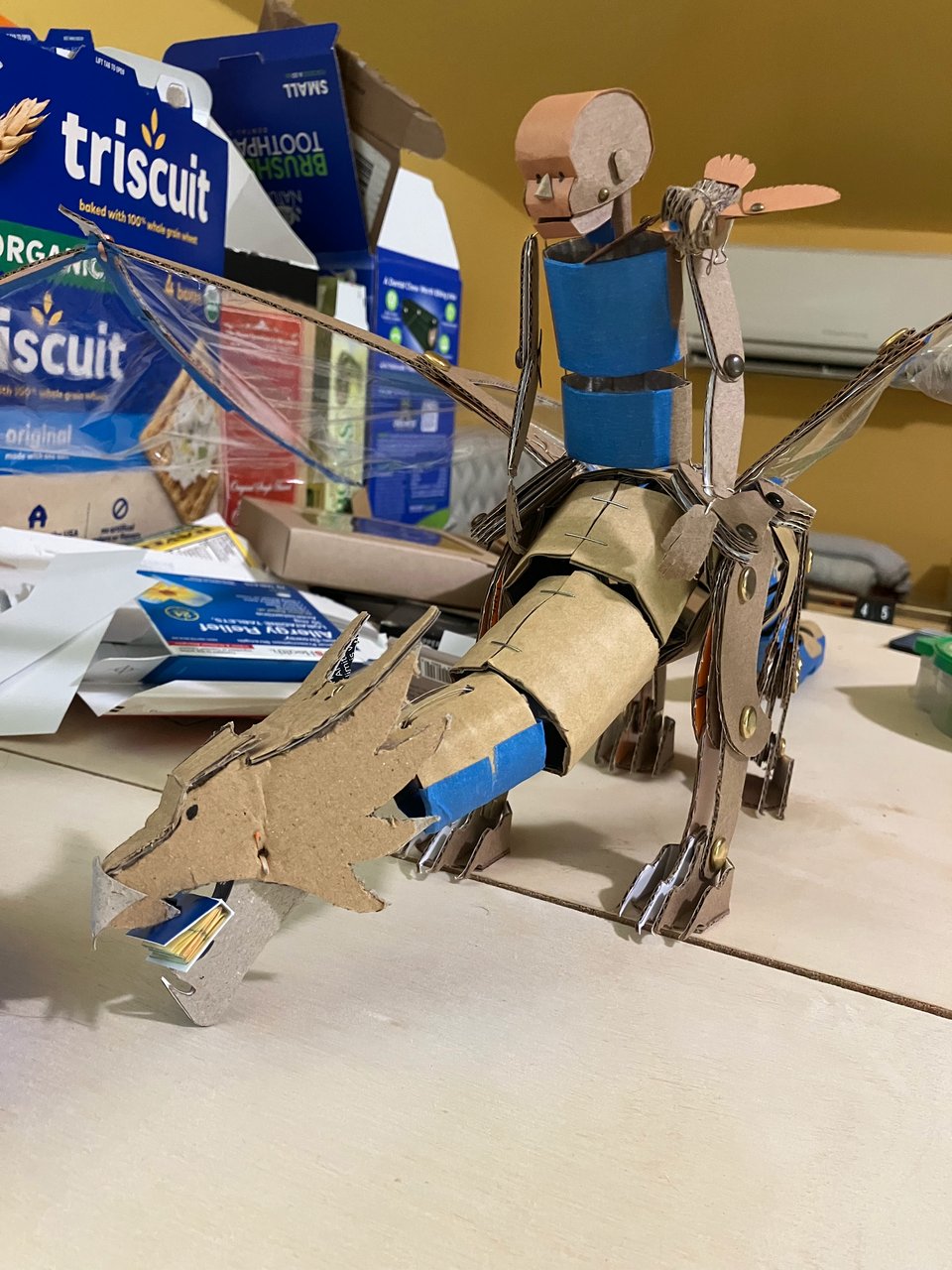 A cardboard puppet of a dragon stands on plywood with a book in its mouth. Atop sits a cardboard puppet of a kid. A cardboard puppet of a (large) bird sits on the kid's shoulder. Behind them is a messy stack of boxes, the most prominent label being "triscuits"