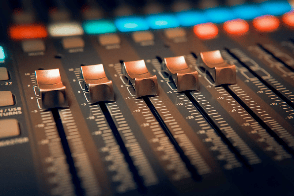A close-up of four slider knobs on a soundboard mixer. Just above the slider knobs you can see a row of brightly lit LED lights, slightly out of focus, mostly blue but a few in white, red, green, and orange.