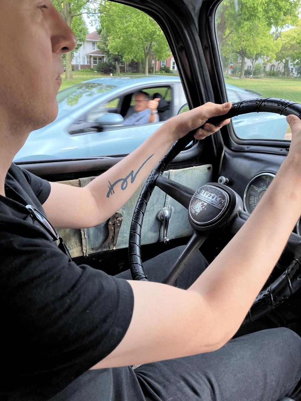 A man drives an old 1950s truck. Through the window you can see another driver flashing a thumbs up