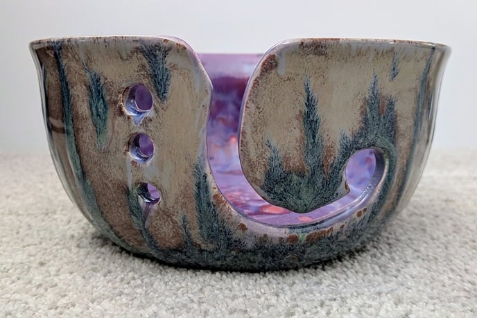 a yarn bowl with three holes in the side and a spiral "cutout" for yarn to be threaded through. It's mottled purple and pink on the interior, and the exterior is a grey/brown with feathery streaks of a muted blue/green, almost like flames or trees in the mis.