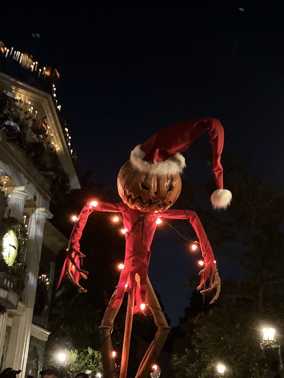 Jack Skellington pumpkin outfit decked in red and hanging on a pole outside the Haunted Mansion in Disneyland. The Jack-O-Lantern head has a grimace and is wearing a Santa hat
