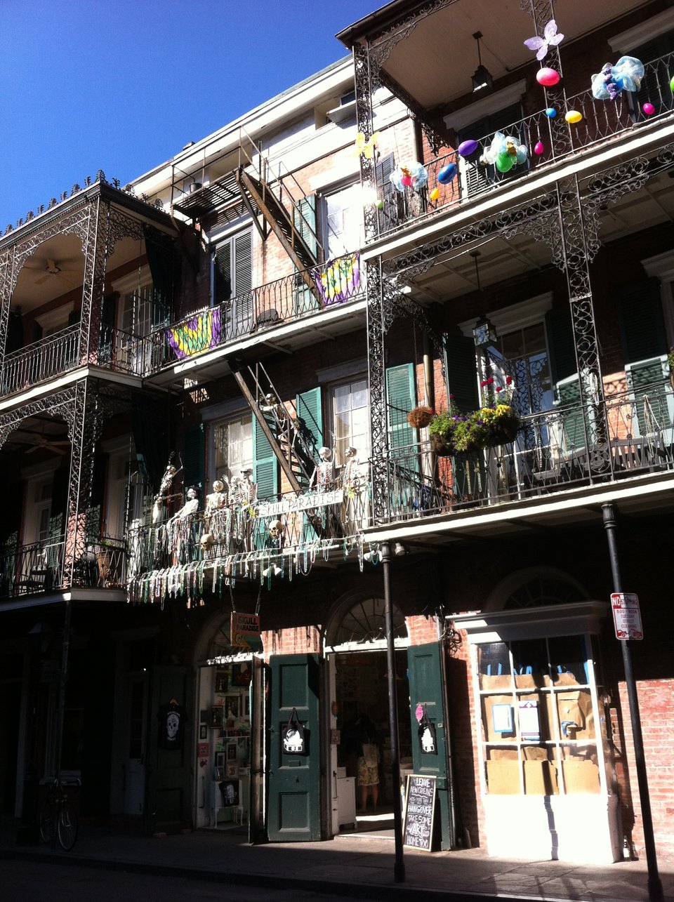 Three-storey buildings on a historic New Orleans street, with shutters and balconies. Balloons hang here and there. In the middle are a bunch of white skeletons lounging on a balcony titled “SKULL PARADISE.”