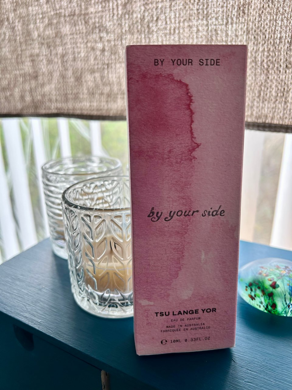 A slim, tall cardboard box in marbled shades of rosy pink is on a dark teal wooden box by a window. The box has By Your Side printed on it. Image by Rowan Ambrose.