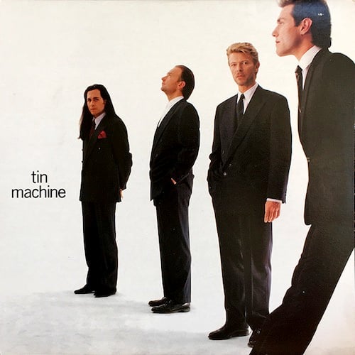 A photograph of the four members of Tin Machine, three white men and David Bowie, who is an alien. They are all dressed in spiffy suits and ties. They are lined up facing to the left, with one member closer to the camera and the last one furthest away from the camera. David Bowie and the guy furthest away from the camera, who has very long brown hair, are looking at the camera while the other two seem to be looking at something to the left and up out of frame.