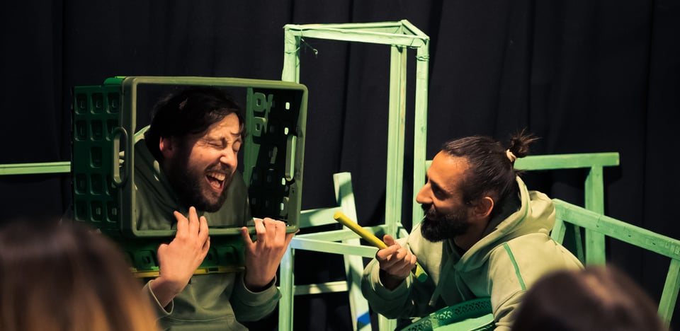 Two persons (Diego Ingold and Gael Doorneweerd-Perry) dressed in green and laughing on stage, amongst green objects.