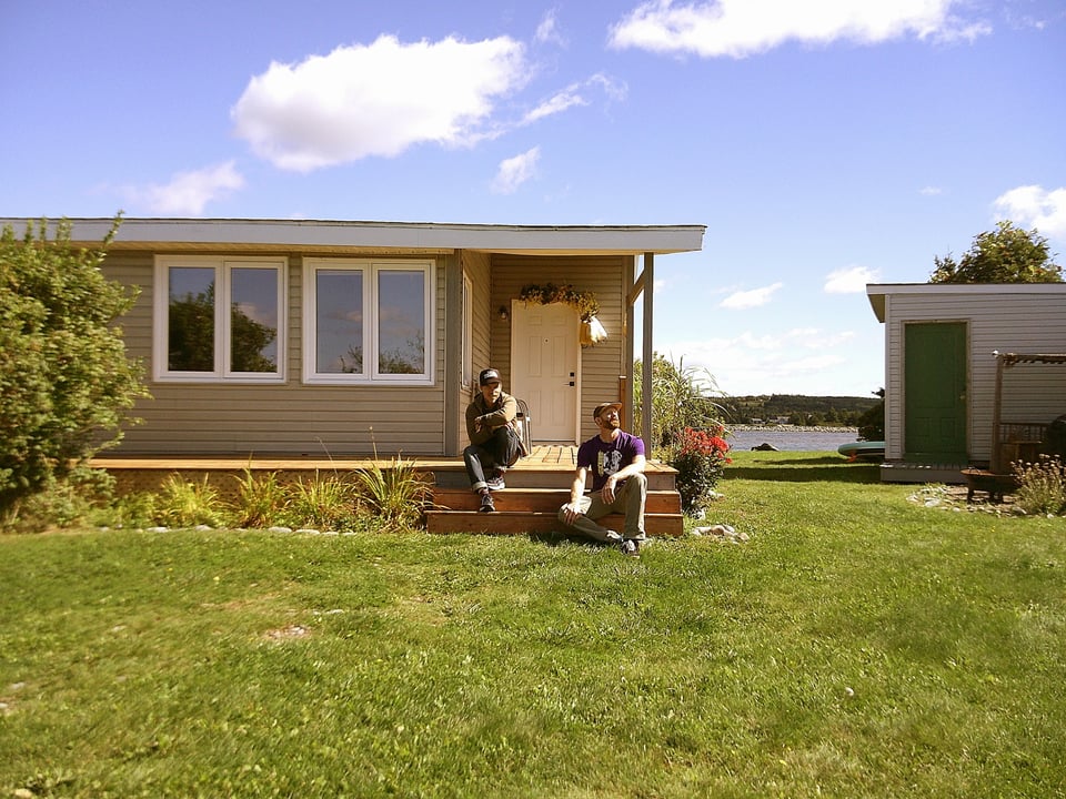 shane and dave sit on the steps in front of a small chalet its a sunny day