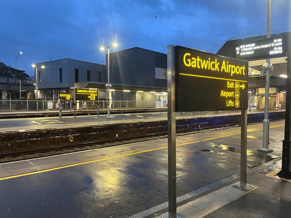 The railway station at Gatwick Airport at night, with a dark blue sky in the backdrop and empty platforms. There’s water on the ground which casts all sorts of reflections.