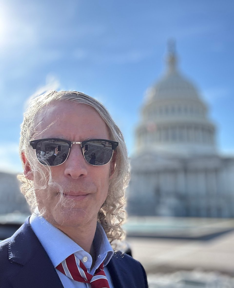 selfie of white man with long hair in a navy suit, light-blue shirt and red/blue and sunglasses, standing in front of the US Capitol building