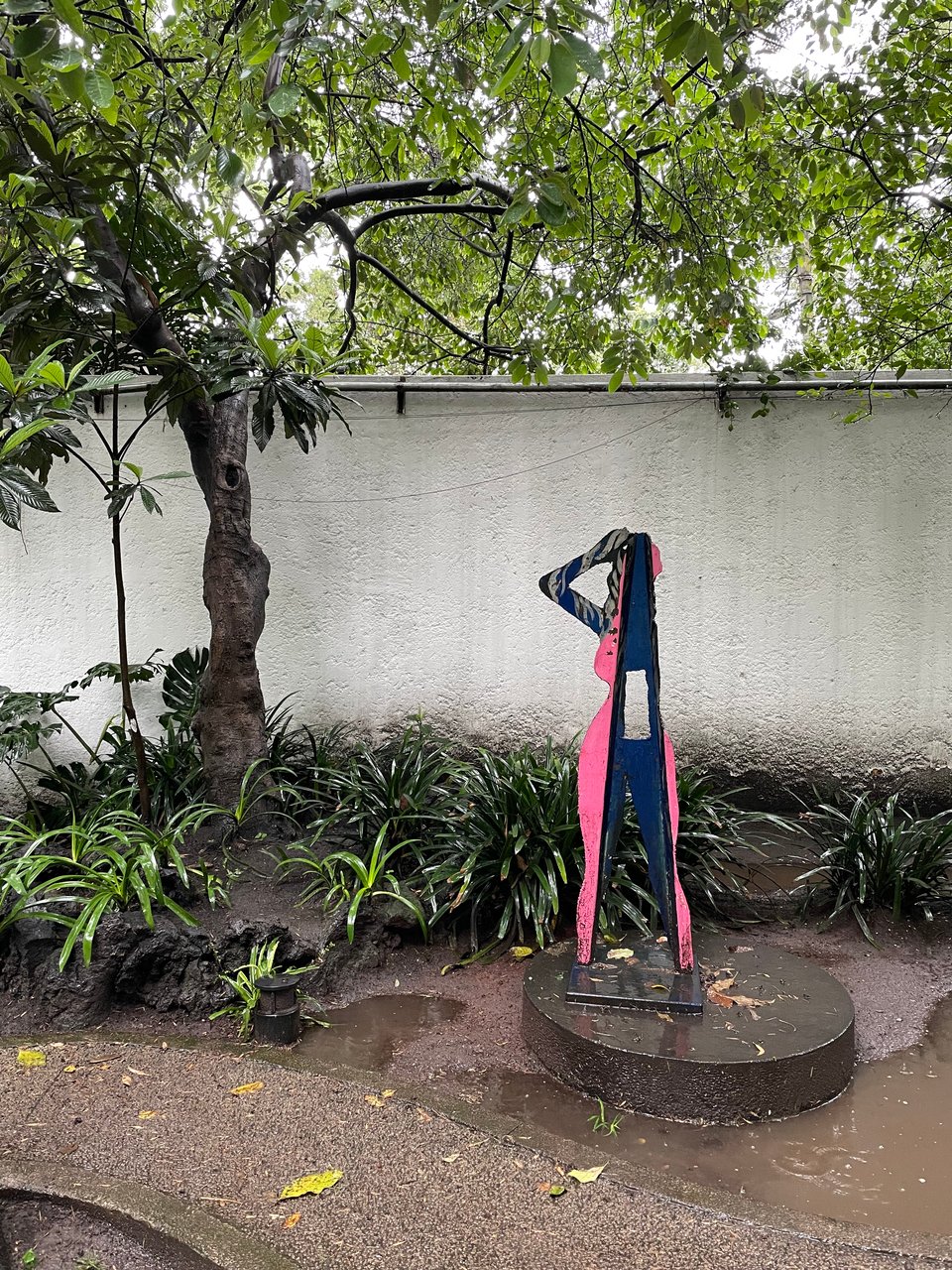 Sculpture of a person in three parts, on a circular pedestal on front of a white garden wall. Alternating blue and pink, blue, and pink.
