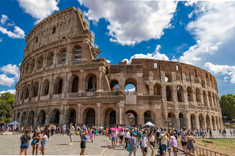 A wide daytime view of the Colosseum in Rome, Italy. The ancient stone amphitheater curves across the frame under a blue sky with scattered clouds. Large crowds of tourists gather around the base, some taking photos and others walking past, emphasizing the monument’s scale.
