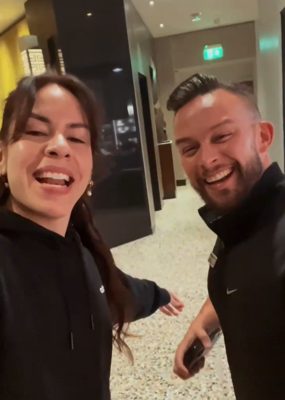 Two people in black tops smile at the camera in a brightly lit hallway. One is holding a phone, and the other has an arm extended.