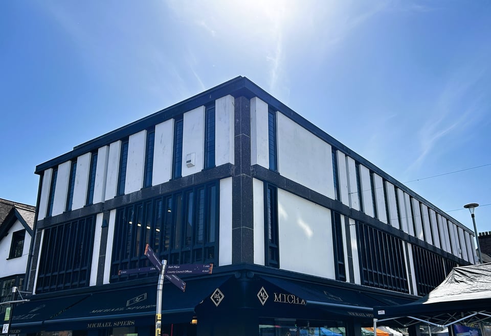 The upper two floors of a shop building on a corner plot. It has a dark concrete frame with panels of white concrete. On the first floor, large windows have been put in put have black vertical frames creating lots of panes. On the second floor, the windows are single slits with dark frames. Overall it looks like a Tudor frame building but with straight lines.