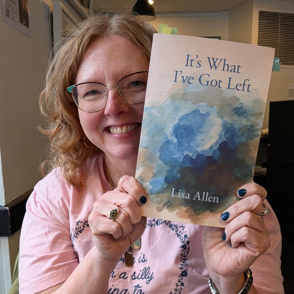 A woman with glasses smiles as she holds her book up for the camera. She's wearing a pink shirt that reads "love is a silly thing to hate." Her book cover is an abstracted blue flower and the title is It's What I've Got Left.