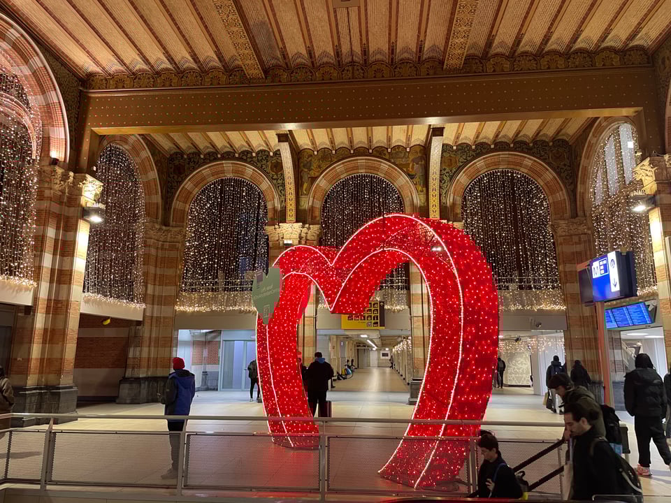 A heart archway inside Amsterdam Centraal train station. There are twinkly lights hanging from the arches of the walls and the heart is covered in red twinkly lights. Very festive scene.