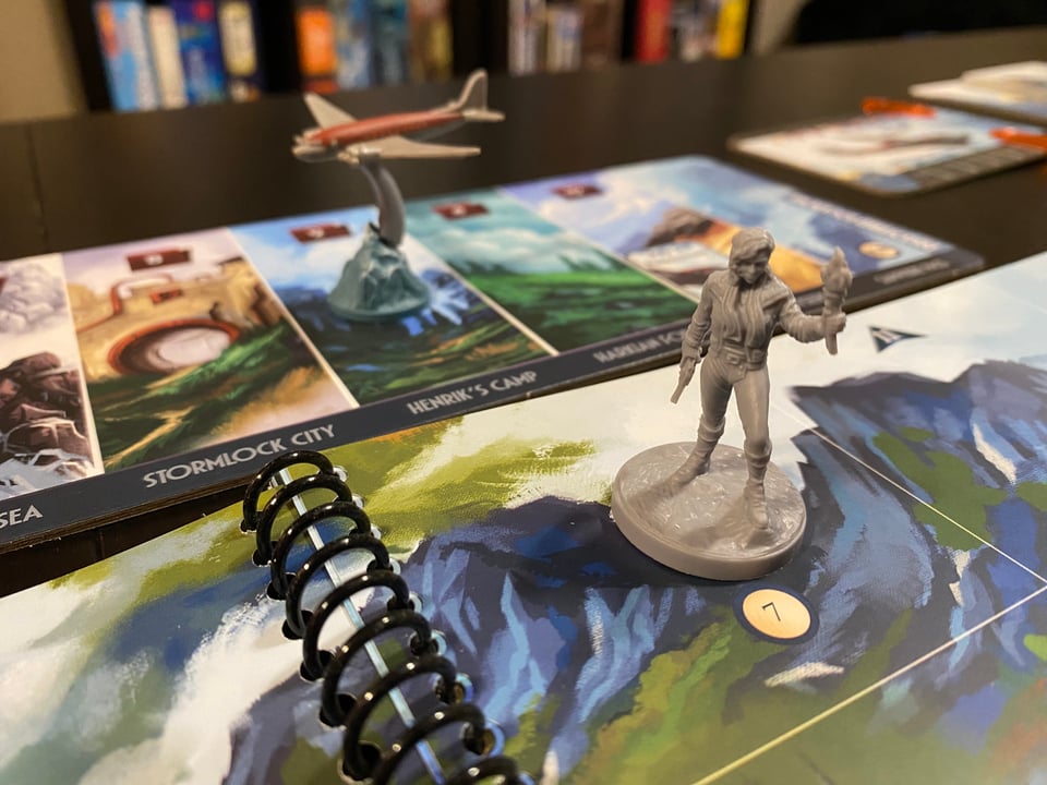 A plastic figurine stands on a map in a spiral bound notebook.  In the background, a plastic airplane stands on a game board.