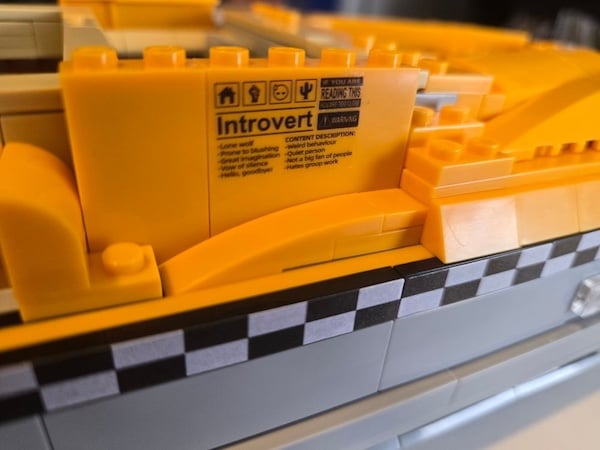A photograph of a Lego piece attached to other Lego pieces and marked with an industrial label that says "Introvert" in Helvetica along with some smaller text that's not really legible, though one line does seem to read "Weird behaviour."