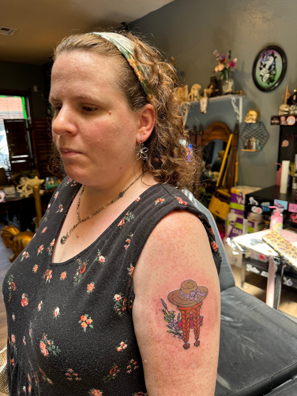 A side view of Daisy, a fair-skinned woman with curly auburn hair held back by a headband, wearing a black floral dress with one sleeve flipped up to display a tattoo on her upper left arm, showing a straw hat with a purple ribbon tied in a bow at the front, 2 bright red-orange braids extending from beneath it, and lavender, violets, and green leaves scattered around the image