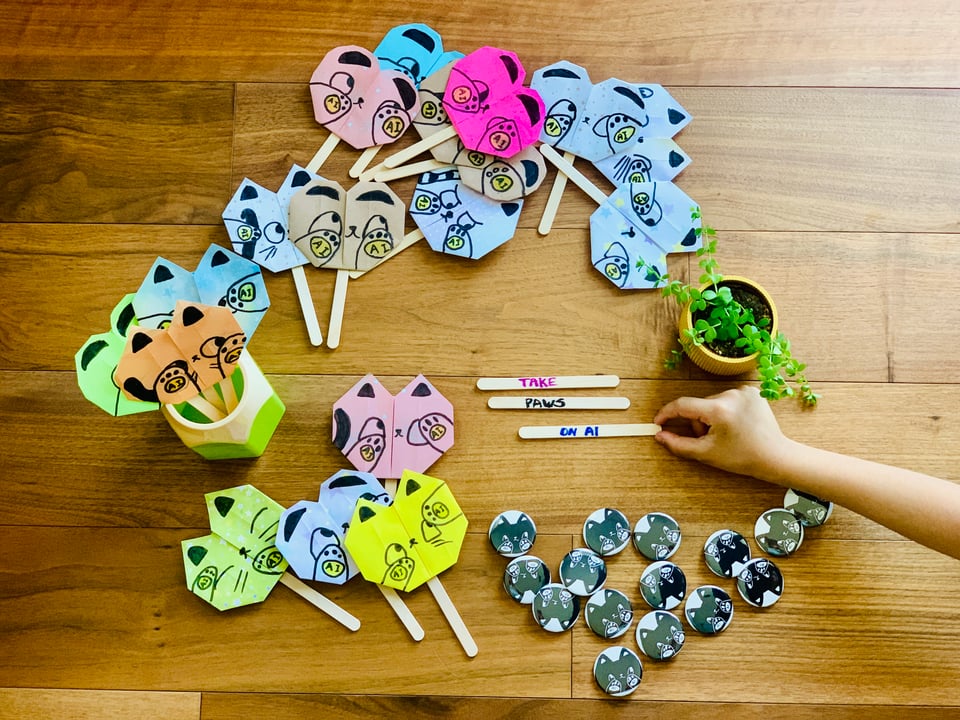 A bunch of cute origami cats attached to popsicle sticks on a hardwood floor. Next to them are some popsicle sticks that say "Take Paws on AI" where pause is spelled like cat paws. A kid is holding one of the popsicle sticks.