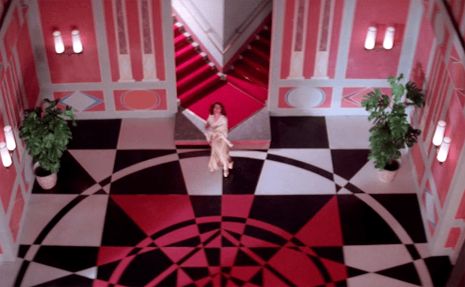 A young woman in a lurid red, white and black Art Deco apartment lobby looks up at the ceiling in Suspiria 1977.