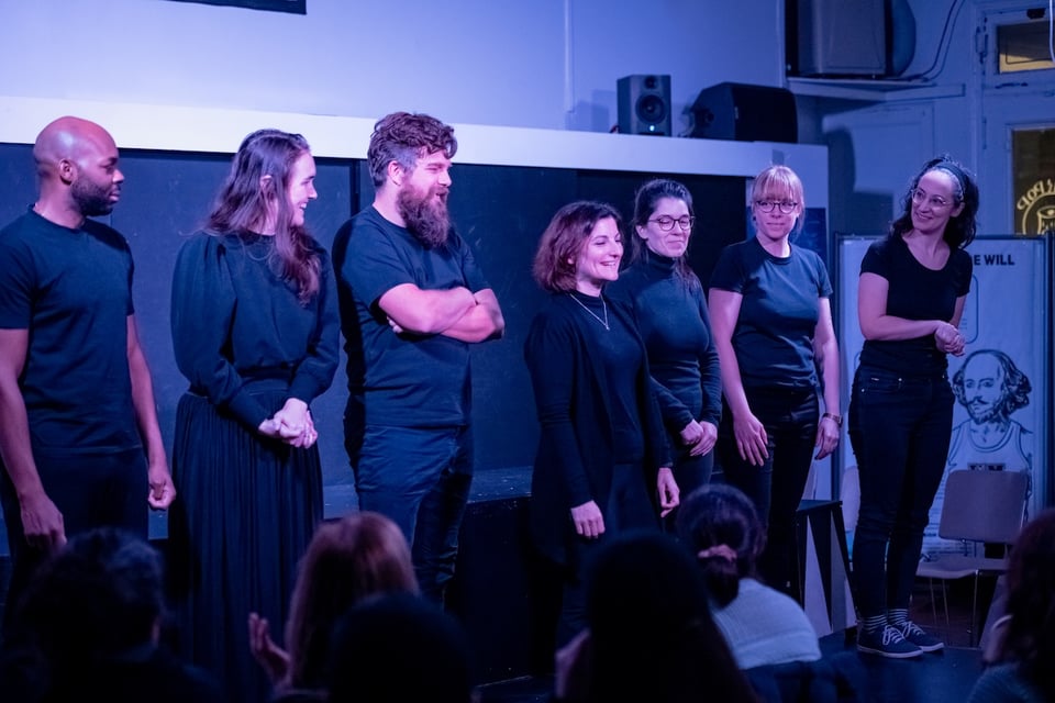 The seven members of the cast of the show standing in a row, all dressed in blalck