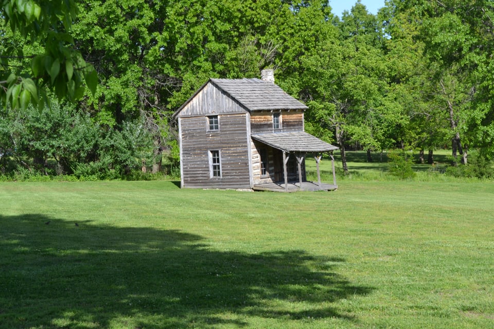 A small, wooden, two storey house with a porch almost as big as it is, surrounded by grass and trees.