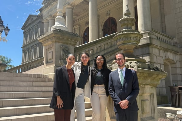 Four people pose together outside the Michigan State Capitol in Lansing on a sunny day.