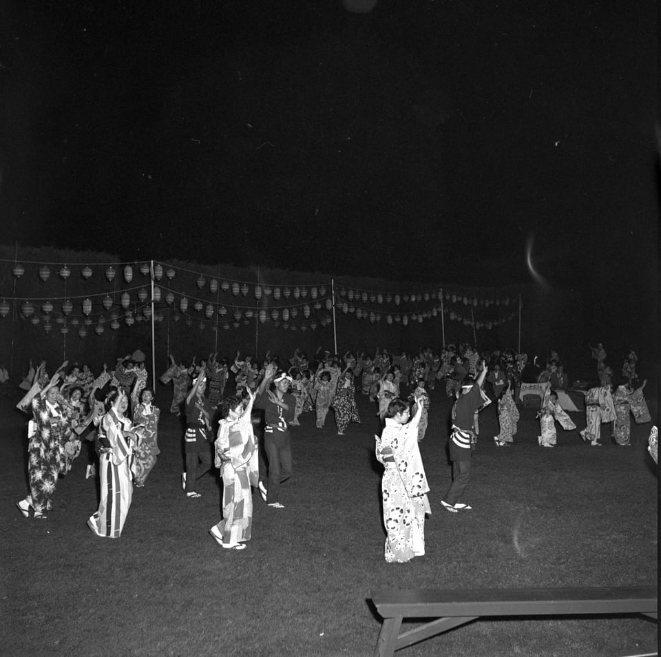 Black and white photograph of a large group of dancers at an Obon festival at night. They are wearing traditional Japanese dress and dancing in unison under a row of hanging lanterns.