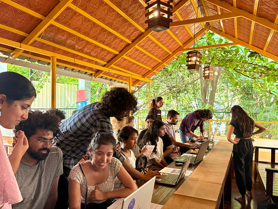 A pop-up lab on a semi-open terrace with a lineup of computers and people engaging with each of those interfaces on one side of the composite table and a table runner print on brown paper on the other side of the same set of tables. The trees in the background and tiny lamps suspended from a gabled roof lend the space its unique brilliance.