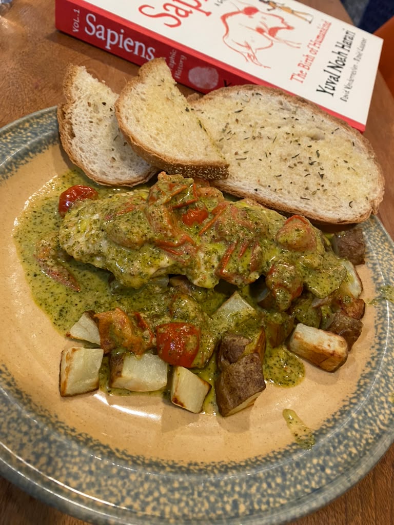 The plated pesto tomato chicken with potatoes and garlic bread. A copy of the graphic novel "Sapiens" is in the background.