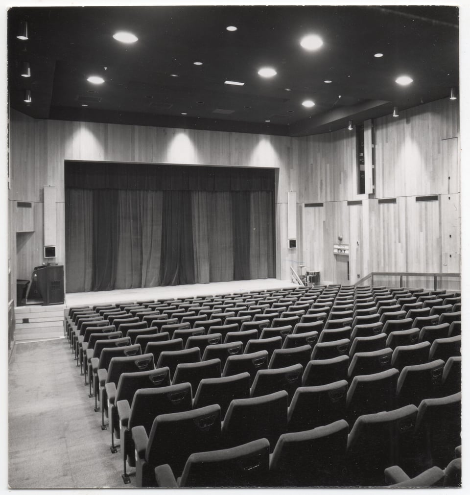 A simple theatre interior, looking towards the stage. There are long rows of seats in a rank (so each row is a little higher than the one before it). The ceiling has recessed house lights. The stage has a very plain and flat proscenium arch.