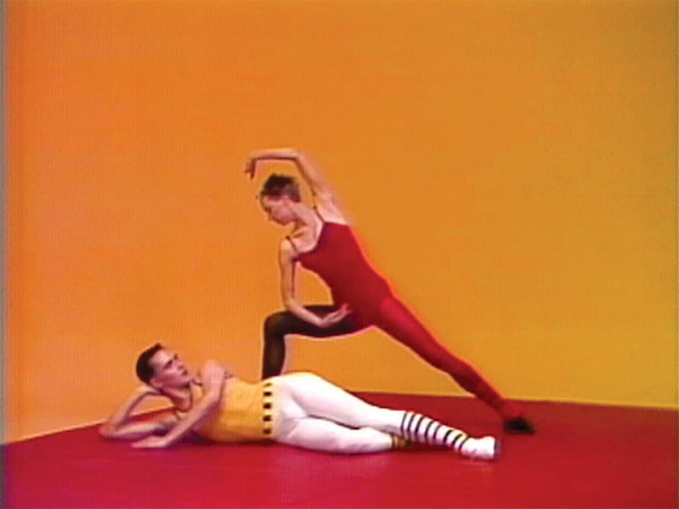 A screenshot of Charles Atlas' Hail The New Puritan. A member of the Michael Clark Company dances over Clark himself, reclining on the floor in patterned outfit.