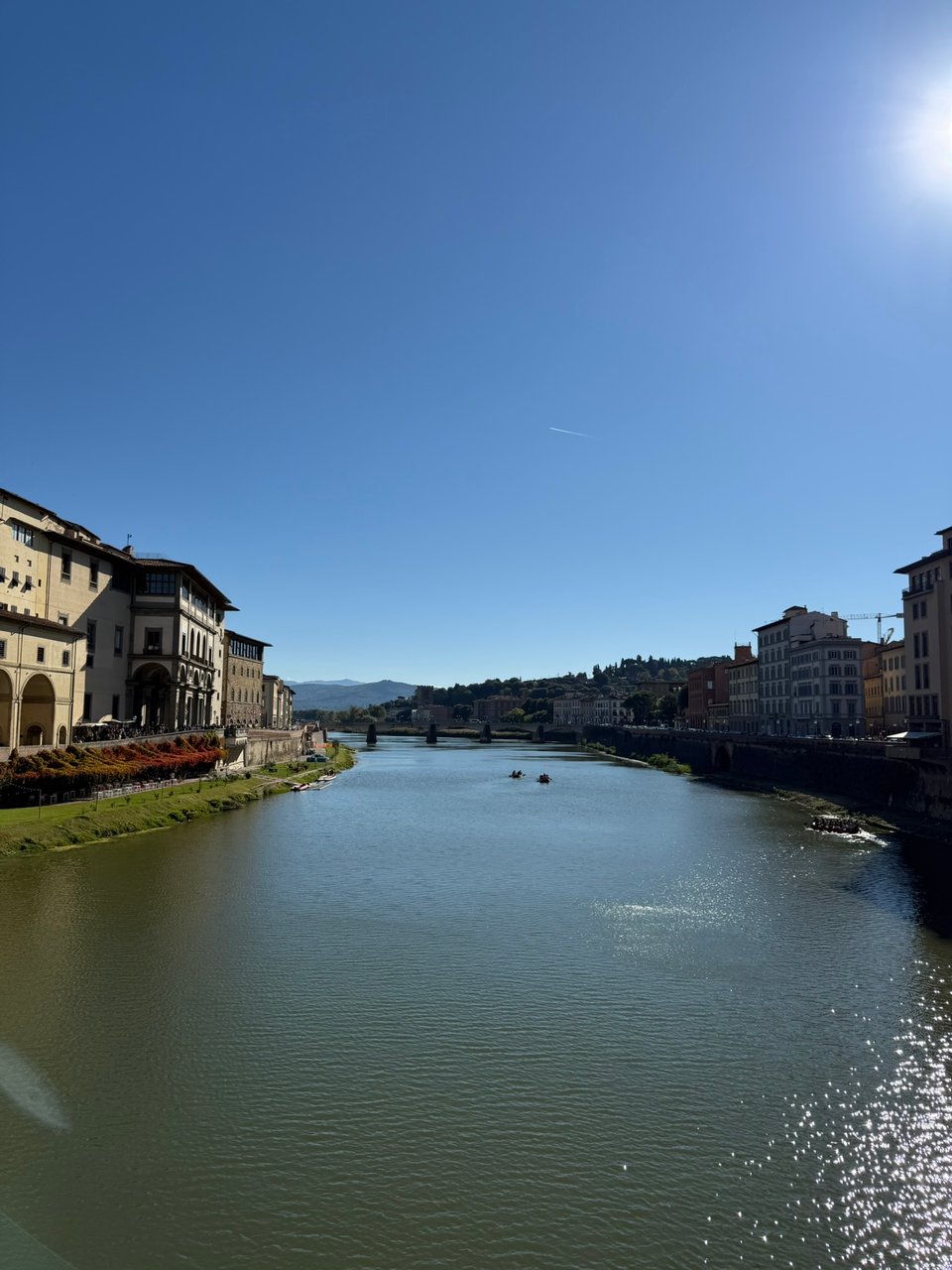 A calm river in Florence flanked by historic buildings on both sides, viewed under a bright, cloudless sky.