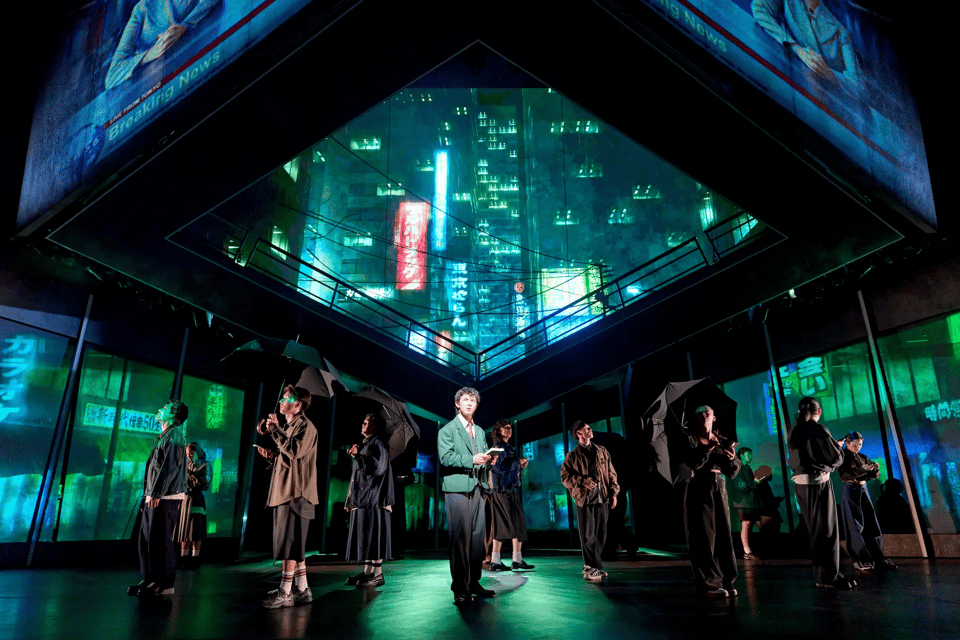 A group of performers in modern attire stand on a stage designed with vibrant, illuminated cityscape projections while holding umbrellas and interacting with electronic devices.