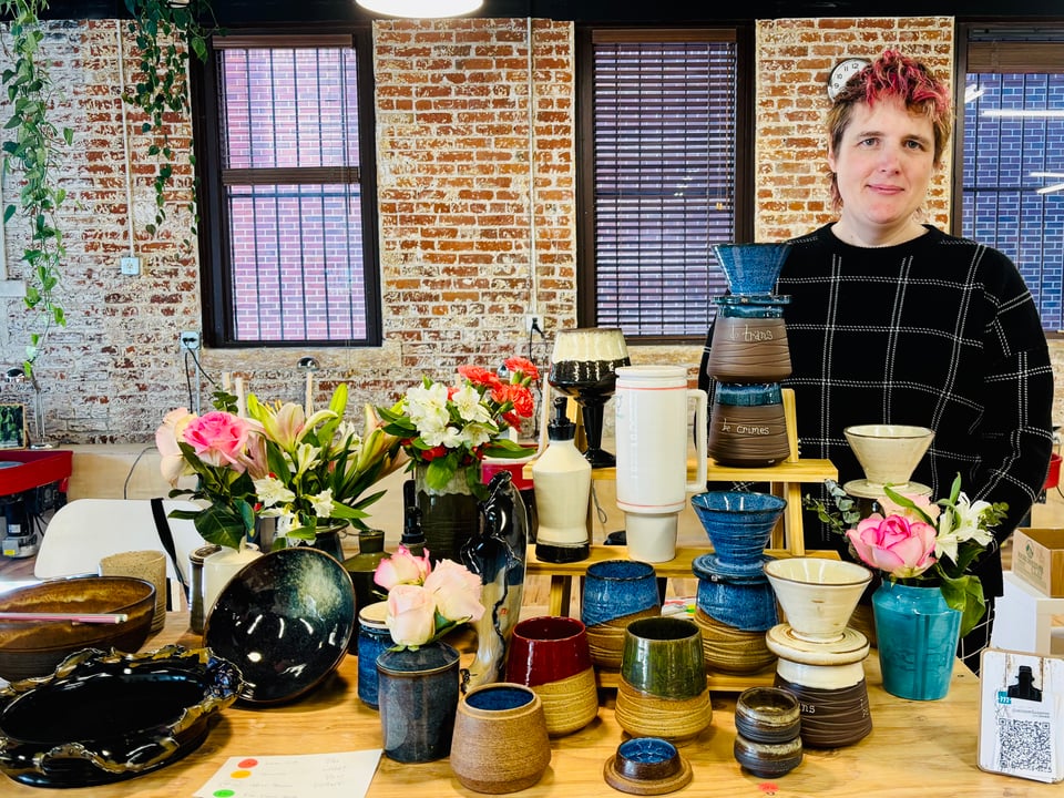 Miriam standing behind a table full of pottery – mugs and jars and vases with flowers and pour-overs and ramen bowls and a large white and blue and pink stanley-cup style lidded vessel