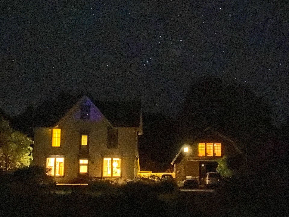 A farm house and a barn side by side are lit up in the night by internal lights. Stars dot the sky overhead, including the Big Dipper which is "upside down".