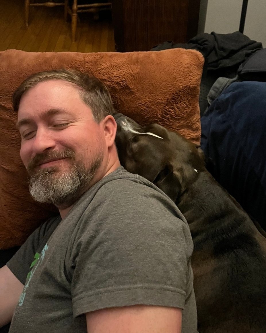 A dark haired pitbull sandwiches herself between her dad (who is laying on the couch) and the couch pillows. Her mouth is right behind his ears.
