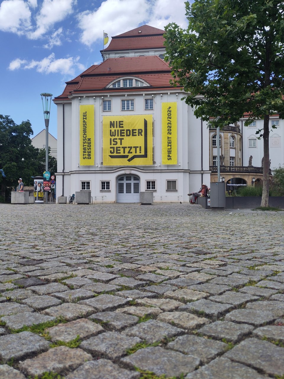 Blick auf eine Hausfassade, darauf banner. Auf einem steht "Staatsschauspiel Dresden", auf einem zweiten "Nie wieder ist jetzt", und auf einem dritten "Spielzeit 2023/2024"